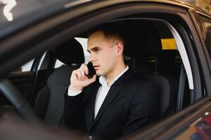 jovem homem de negocios senta dentro luxo carro e fala em telefone. ele parece em linha reta avançar. cara drives carro. ele detém 1 mão em direção roda. isto é ensolarado fora. foto