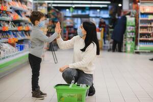 jovem mulher e dela criança vestindo protetora face máscaras fazer compras uma Comida às uma supermercado durante a coronavírus epidemia ou gripe surto. foto