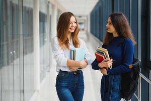 dois jovem mulheres com livro conversando enquanto em pé dentro Faculdade corredor. universidade alunos dentro corredor depois de a palestra. foto