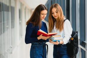 dois jovem mulheres com livro conversando enquanto em pé dentro Faculdade corredor. universidade alunos dentro corredor depois de a palestra. foto