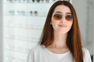 ela fez dela escolha. lindo jovem mulher ajustando dela Novo oculos de sol e sorridente enquanto em pé dentro ótico loja. foto