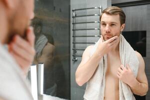 jovem homem olhando dentro espelho depois de barbear às casa foto