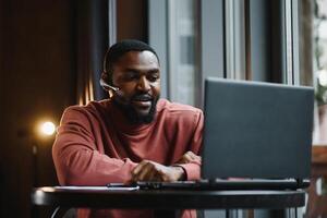 retrato do feliz africano homem de negocios usando telefone enquanto trabalhando em computador portátil dentro restaurante. foto