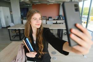 retrato do uma feliz alegre sorridente jovem aluna estudante senhora com grandes cabelo em pé dentro esvaziar Sala de aula olhando Câmera levar uma selfie foto