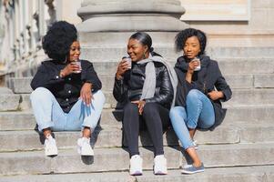 três à moda africano americano meninas bebendo café em a rua e tendo Diversão foto