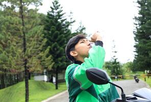 jovem homem moto motorista com conectados motorista aplicação Jaqueta beber uma garrafa do mineral água foto