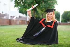 feliz fofa caucasiano grad menina é sorridente. ela é dentro uma Preto argamassa quadro, com vermelho pendão, dentro vestido, com agradável Castanho encaracolado cabelo, diploma dentro mão foto