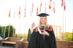 feliz fofa caucasiano grad menina é sorridente. ela é dentro uma Preto argamassa quadro, com vermelho pendão, dentro vestido, com agradável Castanho encaracolado cabelo, diploma dentro mão foto