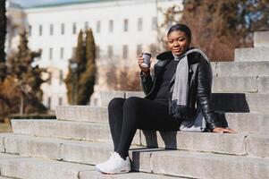 alegre Sombrio esfolado mulher desfrutando café segurando para ir copo recriando dentro cidade parque, feliz na moda vestido africano americano hipster menina sentado ao ar livre durante ensolarado dia bebendo bebida foto