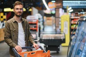 bonito sorridente homem compras dentro supermercado empurrando carrinho foto
