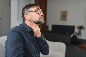 Senior homem sofrimento a partir de frio. foto do idosos homem tendo dolorido garganta. idosos caucasiano homem tocante dele garganta. dolorido garganta, amigdalite ou tireoide glândula problema. mais velho homem às casa sentindo-me indisposto.