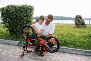 feliz pai e dele filho tendo Diversão juntos às a verde parque, fixação bicicleta junto. do pai dia. foto