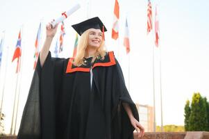 retrato entusiasmado fêmea Faculdade aluna graduado dentro boné e vestido a comemorar, segurando diploma. foto