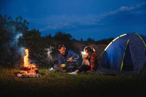 noite acampamento dentro a montanhas. feliz casal Viajantes sentado juntos ao lado fogueira e brilhando turista barraca. em fundo grande pedregulho, floresta e noite céu. foto