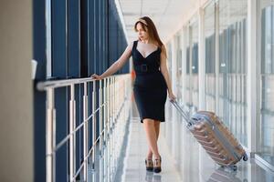 à moda mulher dentro uma Preto vestir com uma mala de viagem dentro a aeroporto terminal esperando para uma avião. foto