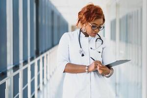 retrato confiante africano americano fêmea médico médico profissional escrevendo paciente notas isolado em hospital clínica corredor janelas fundo. positivo face expressão foto