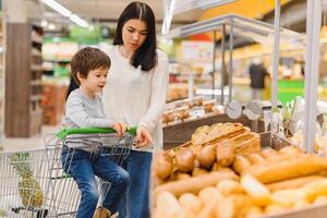 fofa criança com mãe escolhendo cozido bens ou pastelaria dentro padaria fazer compras. pequeno Garoto com mãe comprando pão ou pães às mercearia supermercado foto