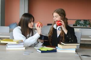 dois feliz sorridente fêmea alunos estão sentado e comendo maçãs, estudando e preparando para exames. foto