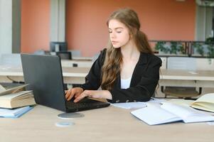 uma lindo caucasiano fêmea aluna é estudando dentro Faculdade remotamente. ela é sentado com uma computador portátil e uma bloco de anotações e concentrado é assistindo uma vídeo conferência lição foto