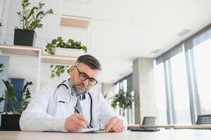 Senior doutor, escrevendo e retrato do uma hospital trabalhador dentro uma escritório fazendo médico pesquisar. felicidade, computador portátil e lista de controle do uma bem estar e saúde empregado com uma sorrir a partir de clínica visão foto