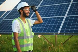 a indiano masculino engenheiro dentro uma verde colete é trabalhando em uma campo do solar painéis. a conceito do renovável energia. foto