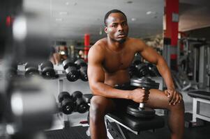jovem africano americano homem sentado e elevação uma haltere fechar para a prateleira às academia. masculino peso Treinamento pessoa fazendo uma bíceps ondulação dentro ginástica Centro foto
