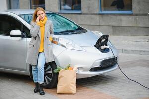 tendo uma conversa usando o telefone. mulher na estação de carga de carros elétricos durante o dia. veículo novo foto