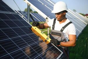 afro-americano trabalhador dentro cinzento macacão e uma branco Difícil chapéu trabalho dentro uma campo do solar painéis. solar renovável energia conceito. foto