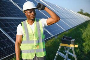 africano americano homem dentro branco capacete e cinzento macacão em pé entre linhas do solar painéis foto