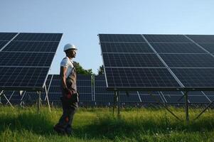 competente energia engenheiro dentro cinzento macacão e branco capacete verificação solar painéis enquanto caminhando em campo. africano americano homem carregando prancheta e recipiente com instrumentos foto