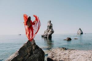 mulher viagem mar. jovem feliz mulher dentro uma grandes vermelho vestir posando em uma de praia perto a mar em fundo do vulcânico rochas, gostar dentro Islândia, partilha viagem aventura viagem foto