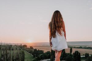 feliz mulher em pé com dela costas em a pôr do sol dentro natureza dentro verão com aberto mãos. romântico lindo noiva dentro branco boho vestir posando com montanhas em pôr do sol foto