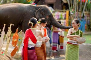 fechar-se lindo tailandês jovem senhora porcelana tailandês tradicional vestir usar flores para borrifar água em cada de outros em a tailandês Novo anos dia dentro uma Diversão caminho em borrado elefante e pilha do areia fundo. foto