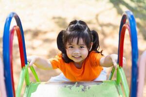 uma jovem menina é sorridente Como ela sobe uma verde e azul Parque infantil deslizar. a deslizar é localizado dentro uma parque, e a menina é vestindo a laranja camisa foto