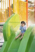 uma pequeno menina é sentado em uma verde deslizar às uma água parque. ela é vestindo uma azul e branco roupa de banho e é olhando baixa às dela face foto