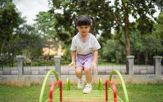 ásia pequeno menina é jogando em uma verde e vermelho Parque infantil estrutura. ela é vestindo uma branco camisa e roxa calção foto
