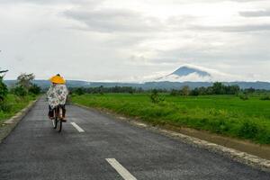 ásia agricultor mulher equitação uma bicicleta às a manhã em a asfalto estrada. ciclismo com montanha e arroz arroz campo Visão às a fundo. cópia de espaço foto