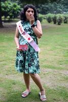 uma grávida indiano senhora poses para ao ar livre gravidez tiro e mãos em barriga, indiano grávida mulher coloca dela mão em dela estômago com uma maternidade vestir às sociedade parque, grávida lado de fora maternidade tiro foto