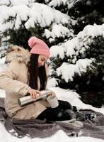 Mulher bonita em roupas quentes de inverno segurando uma garrafa térmica e bebendo chá ou café quente ao ar livre em dia de neve foto