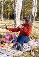 Mulher bonita em um suéter vermelho em um piquenique em uma floresta de outono usando dispositivos móveis