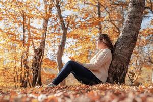 jovem mulher pensativa sentada perto de uma árvore na floresta de outono foto