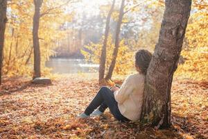 jovem mulher pensativa sentada perto de uma árvore na floresta de outono foto