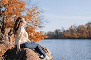 mulher romântica e atenciosa sentada nas rochas na margem do rio no pôr do sol no dia de outono foto
