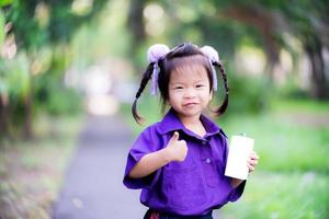 menina asiática estudante segurando uma caixa de papelão de leite branco com canudo azul. criança segurando a caixa de suco de frutas com a mão esquerda. criança mostra o polegar e um sorriso doce. as crianças usam uniforme escolar roxo. 3 anos de idade. foto