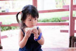 adorável garota asiática comendo pão durante a viagem. criança bonitinha está com fome. as crianças usam lindas roupas escuras. criança tem 3 anos. copie o espaço.