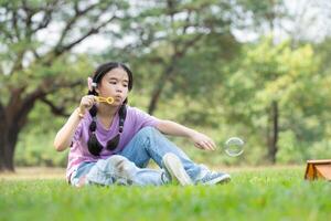 menina sentado dentro a parque com sopro ar bolha, cercado de vegetação e natureza foto