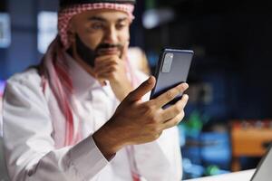 uma saudável árabe homem de negocios usando uma Móvel telefone às dele escrivaninha. detalhado imagem do uma muçulmano cara surfar a líquido, conduzindo pesquisar, e comunicando conectados com dele Smartphone foto