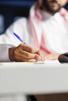 uma focado homem de negocios do meio Oriental etnia eficientemente trabalho às dele casa escritório mesa, levando pesquisa notas usando uma azul caneta. seletivo foco a árabe pessoa escrevendo baixa em dele caderno. foto