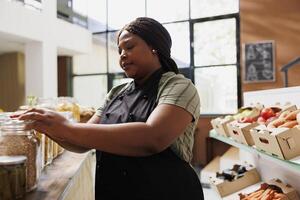 jovem africano americano mulher comerciante dentro uma pequeno sustentável loja, oferta localmente crescido e livre de produtos químicos produtos para eco-consciente clientes. fornecedor organizando frascos preenchidas com orgânico bio Unid. foto