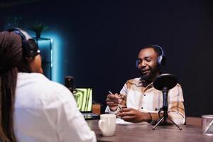 africano americano casal vestindo sem fio fones de ouvido estão fazer uma podcast às casa usando audio gadgets. Preto masculino hospedeiro entrevistando fêmea blogueiro para dele conversa mostrar conectados público. foto